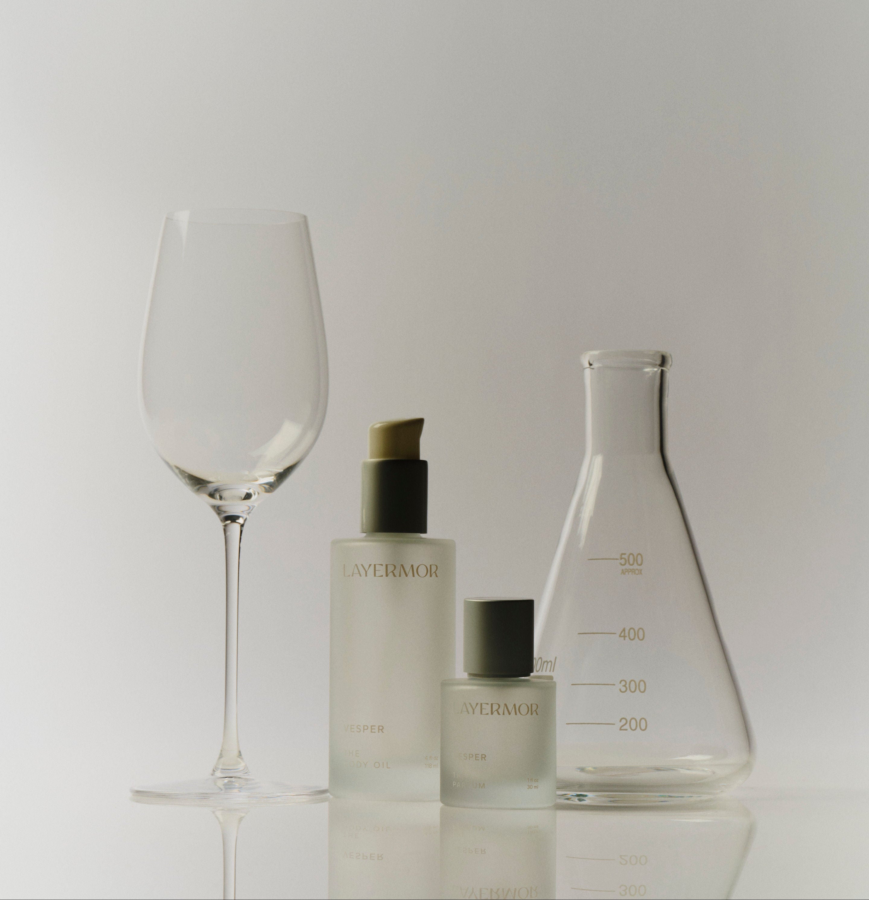 Three frosted glass laboratory-style containers and a clear wine glass on a reflective white surface against a light gray background. The containers show the Lavermook brand name in gold lettering.