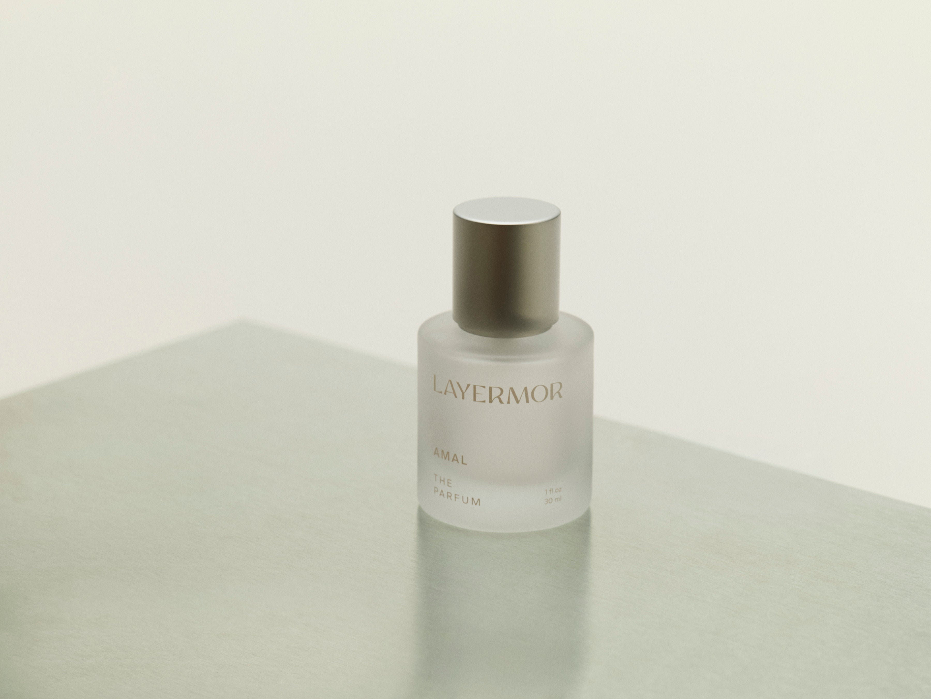 A frosted glass bottle of LAYERMOR The Parfum with a metallic silver cap, standing on a light gray surface against a warm off-white background.