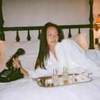 A woman in a white robe reclines in bed with white linens, holding a black cordless phone. A silver tray with two clear bottles sits on the bed in front of her.